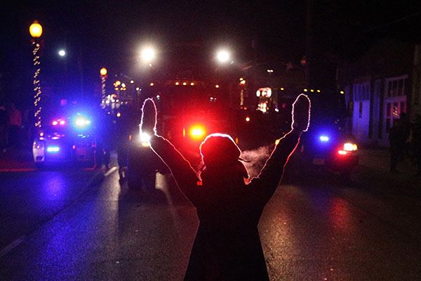 A protester raises her hands in the street as police use tear gas to try to take control of the scene near a Ferguson Police Department squad car after protesters lit it on fire.  RBHS was curiously silent through these events.